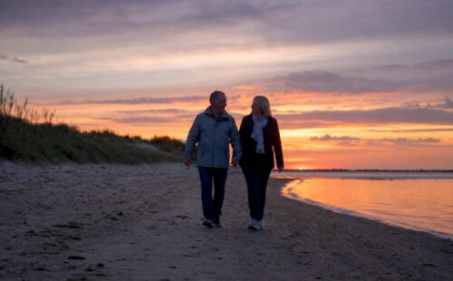 Stelletje wandelt samen over het strand van een Waddeneiland terwijl de zon langzaam ondergaat.