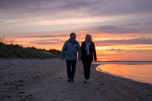 Stelletje wandelt samen over het strand van een Waddeneiland terwijl de zon langzaam ondergaat.